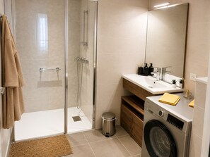 A modern bathroom with a glass shower enclosure, a sink with a mirror, a washing machine, and a trash bin. - Zenao Appart'hôtels Aix Carnot (Aix-en-Provence)