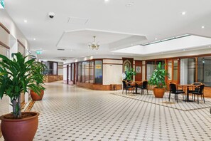 Eine geräumige Hotelhalle mit einem gemusterten Boden, großen Topfpflanzen, einem Kronleuchter und einer Sitzgruppe mit Tischen und Stühlen.