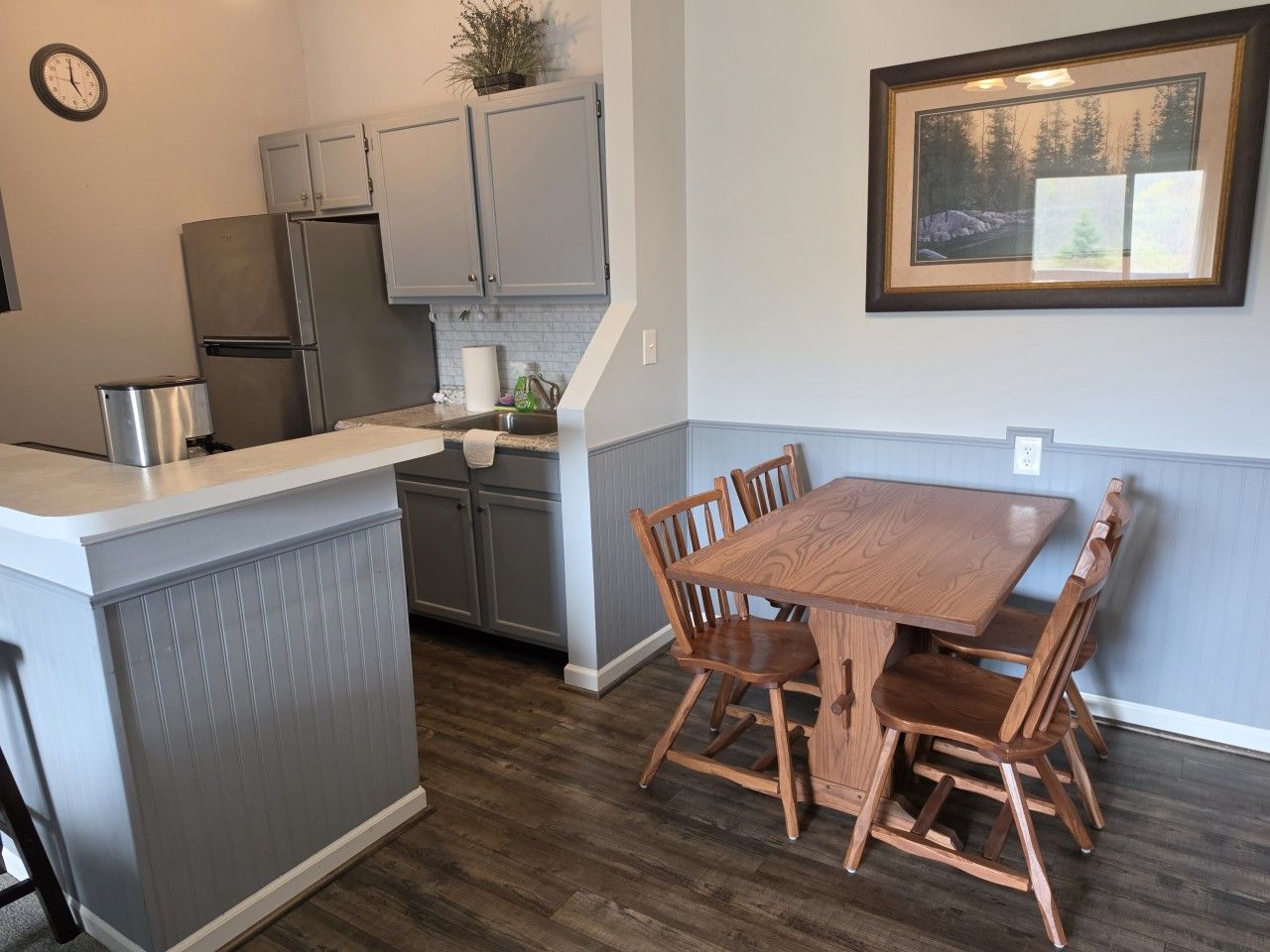 A kitchen with grey cabinets, a stainless steel refrigerator, a wooden dining table with chairs, and a framed picture on the wall.