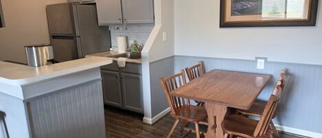 A kitchen with grey cabinets, a stainless steel refrigerator, a wooden dining table with chairs, and a framed picture on the wall.