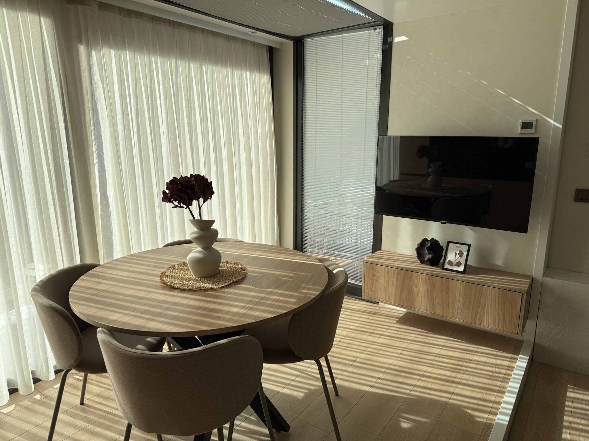 Un comedor moderno con una mesa redonda, cuatro sillas, un jarrón con flores y un televisor montado en la pared.