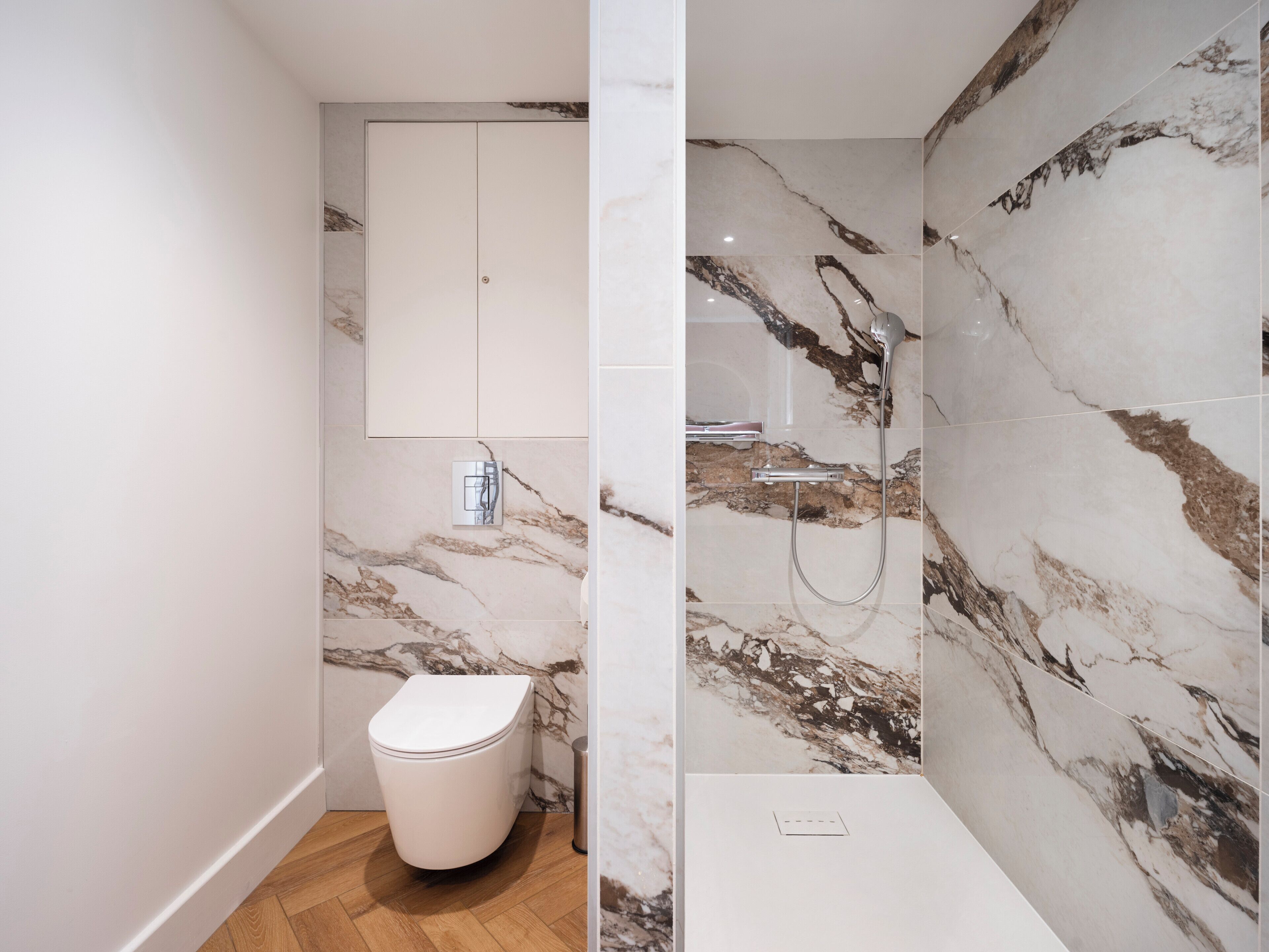 A bathroom with a toilet, a shower with marble walls, and a wooden floor.