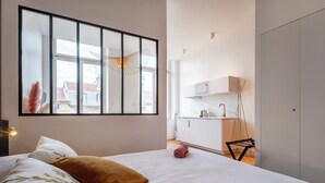 Un dormitorio con un ventanal grande, una cama con ropa de cama floral, una mesita de noche y vistas a una zona de cocina.