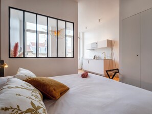 Ein Schlafzimmer mit einem großen Fenster, einem Bett mit floralem Bettbezug, einem Nachttisch und Blick auf einen Küchenbereich.