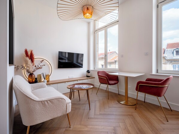 Una sala de estar moderna con un sofá, dos sillones rojos, una mesita redonda, un televisor montado en la pared y un ventanal con vistas a edificios.