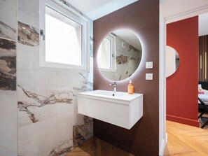 Un baño con pared de mármol, lavamanos blanco, espejo y una botella de jabón.