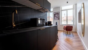 A modern kitchen with dark cabinetry, a black countertop, and a gold faucet. There is a window providing natural light and a view of buildings outside. - Les perchoirs de Stanislas 2 - logement de standing hyper centre (Nancy)
