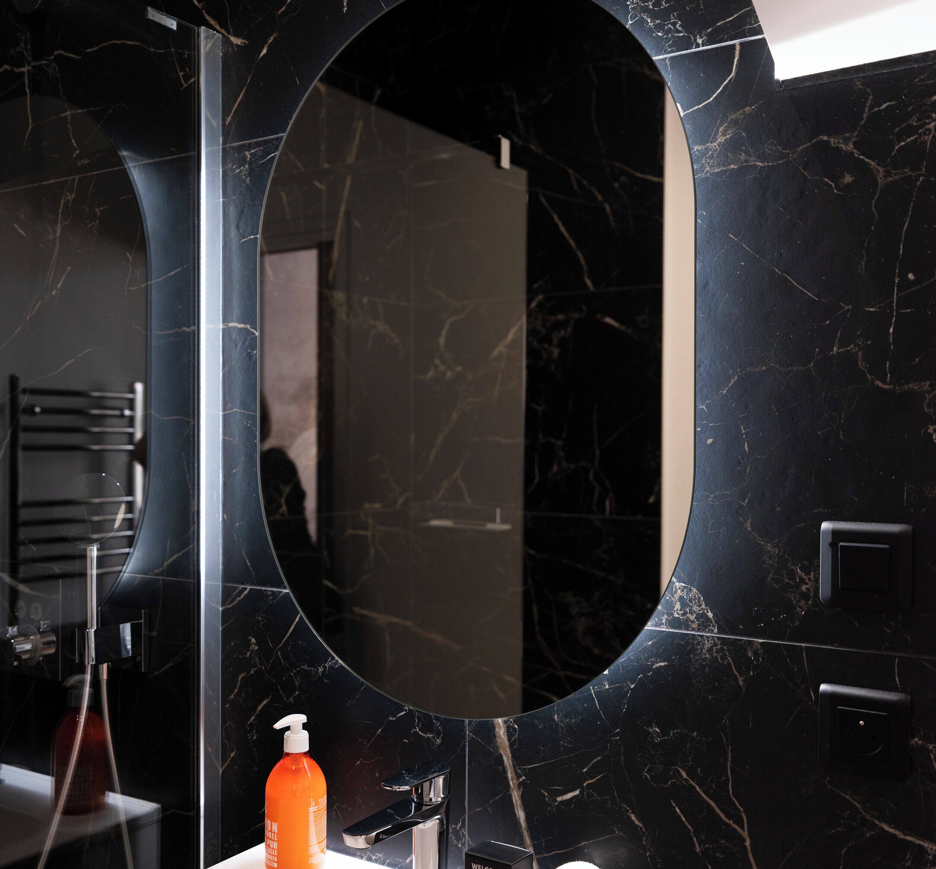 Une salle de bain avec un grand miroir ovale, un lavabo moderne et une bouteille de savon liquide orange.