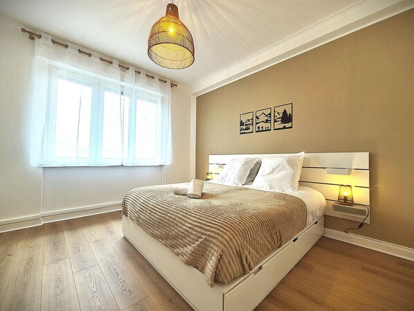 Une chambre à coucher avec un lit, une fenêtre avec des rideaux, une lampe fixée au mur et un luminaire au plafond.