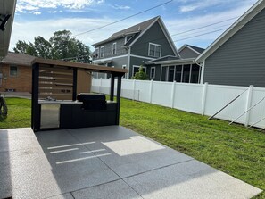 Ein Patio-Bereich mit Grill und Sitzgelegenheit, umgeben von einem weißen Zaun und Häusern im Hintergrund.