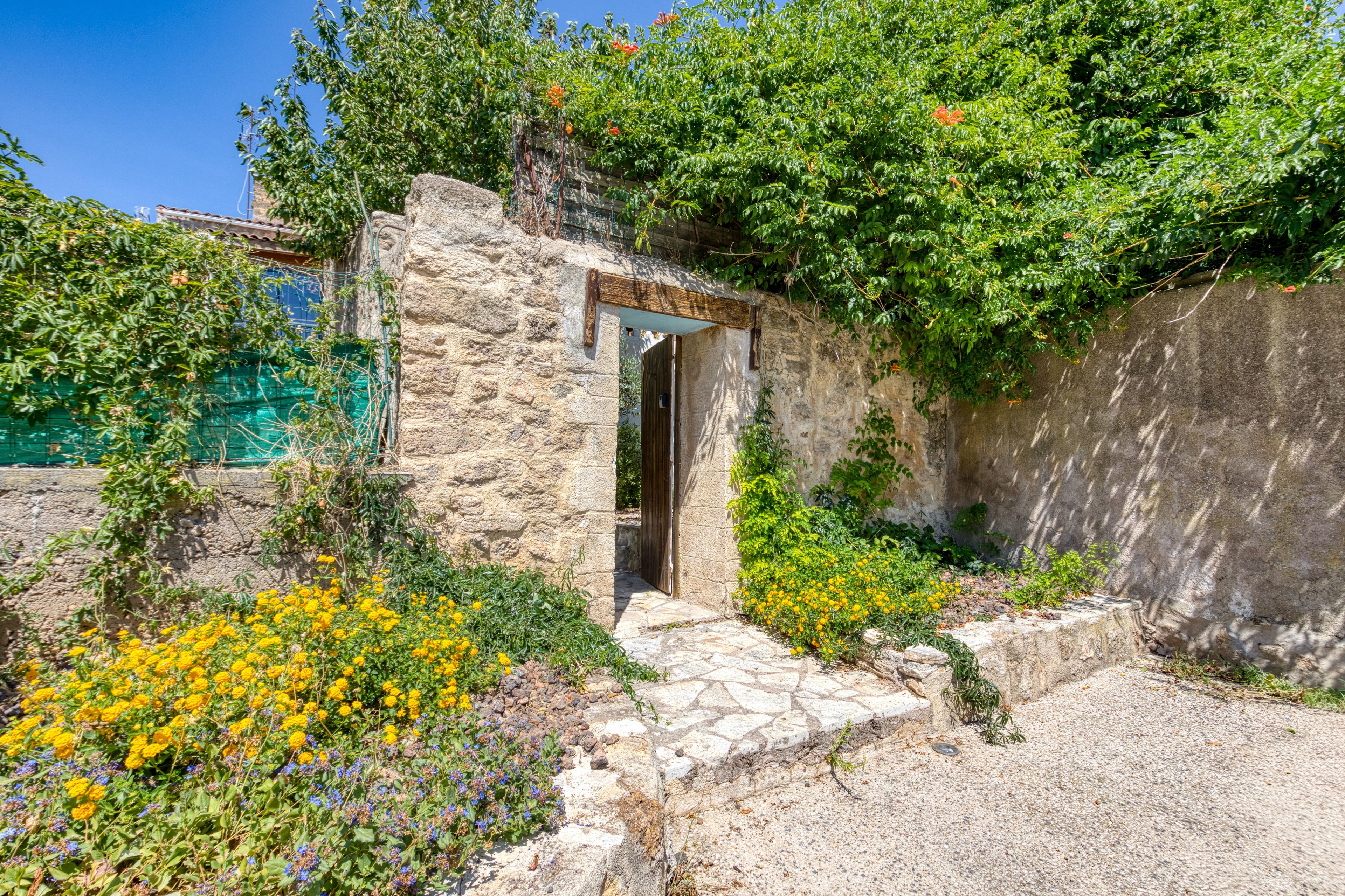 Une construction en pierre avec une porte en bois, entourée de verdure et de fleurs jaunes.