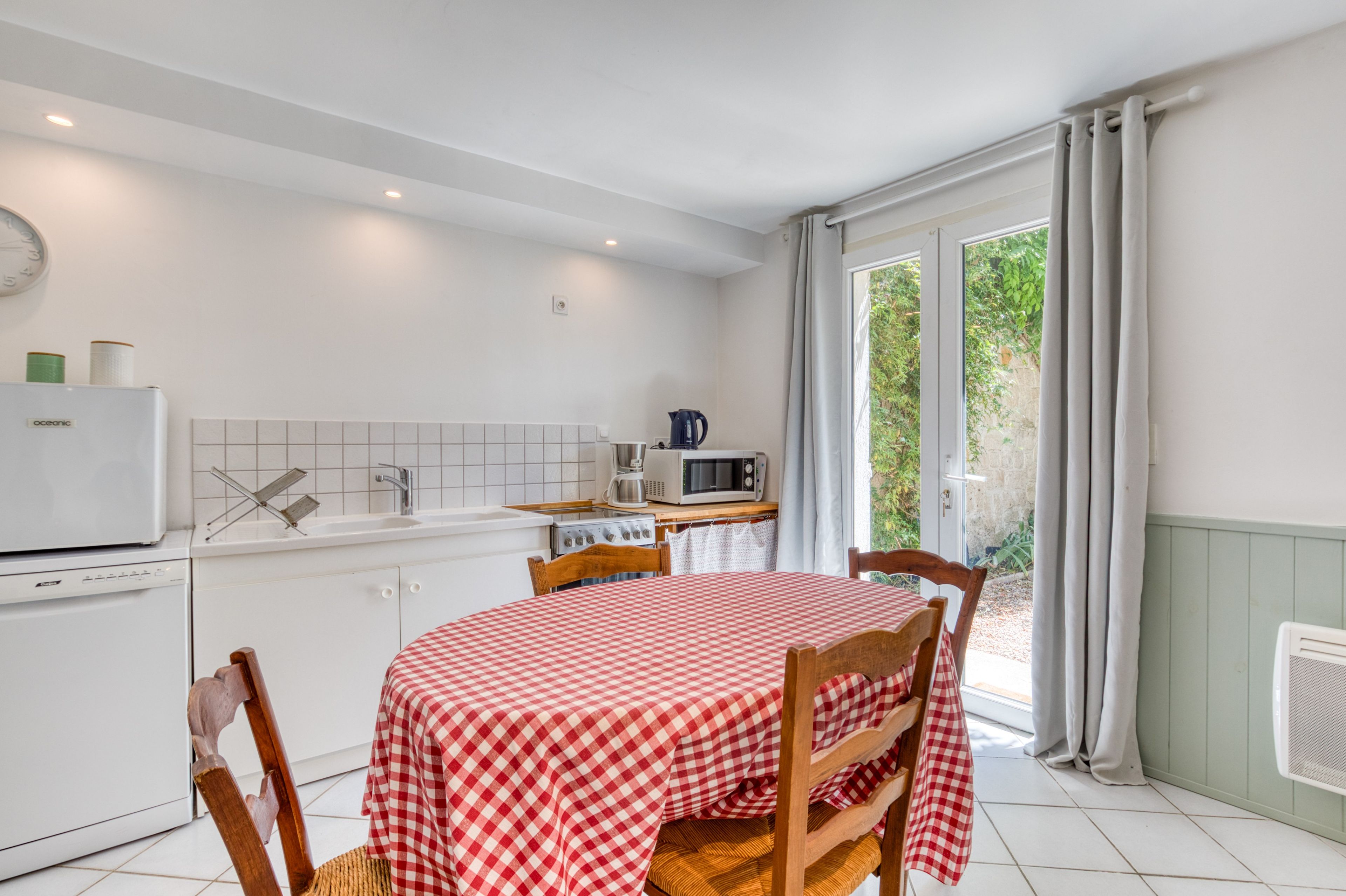 Een keuken met een eettafel, afgedekt met een geruit tafelkleed, houten stoelen, een witte koelkast, een magnetron en een gootsteen.