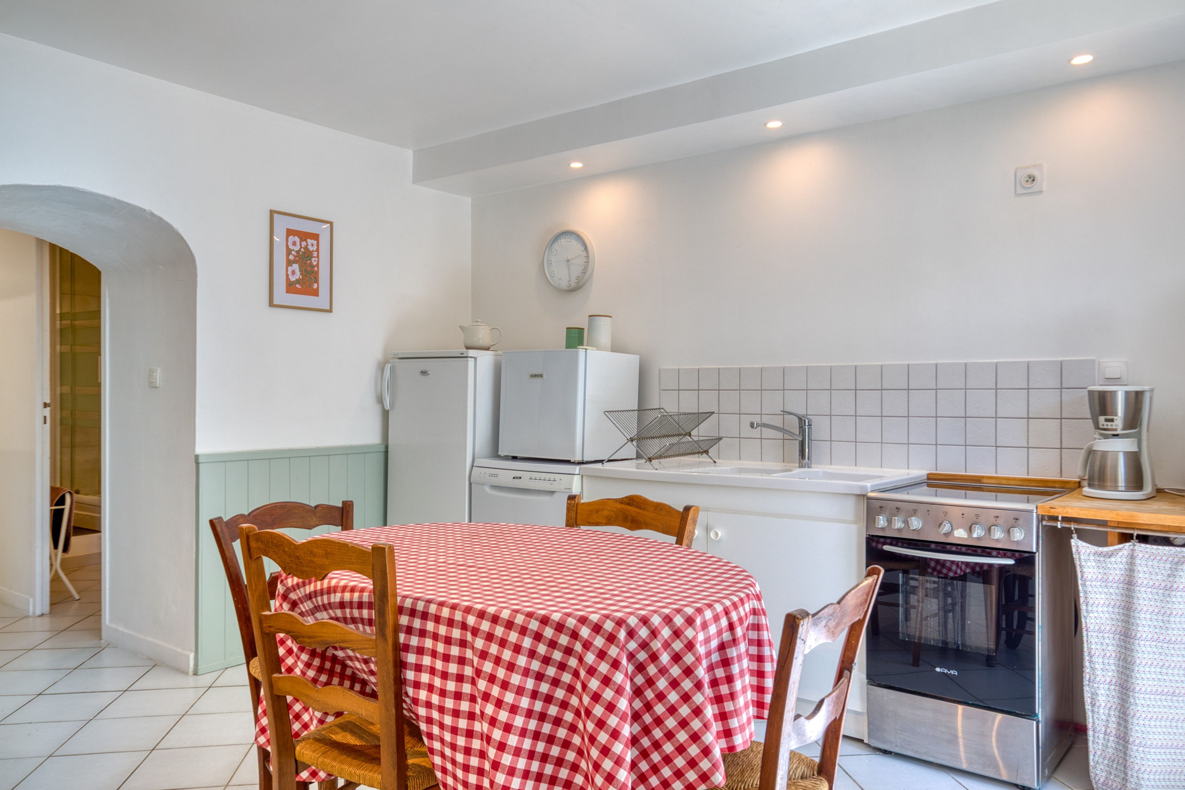 Eine Küche mit kariertem, rot-weißen Tischläufer, Holzstühlen, einem Kühlschrank, einem Herd und einer Uhr an der Wand.