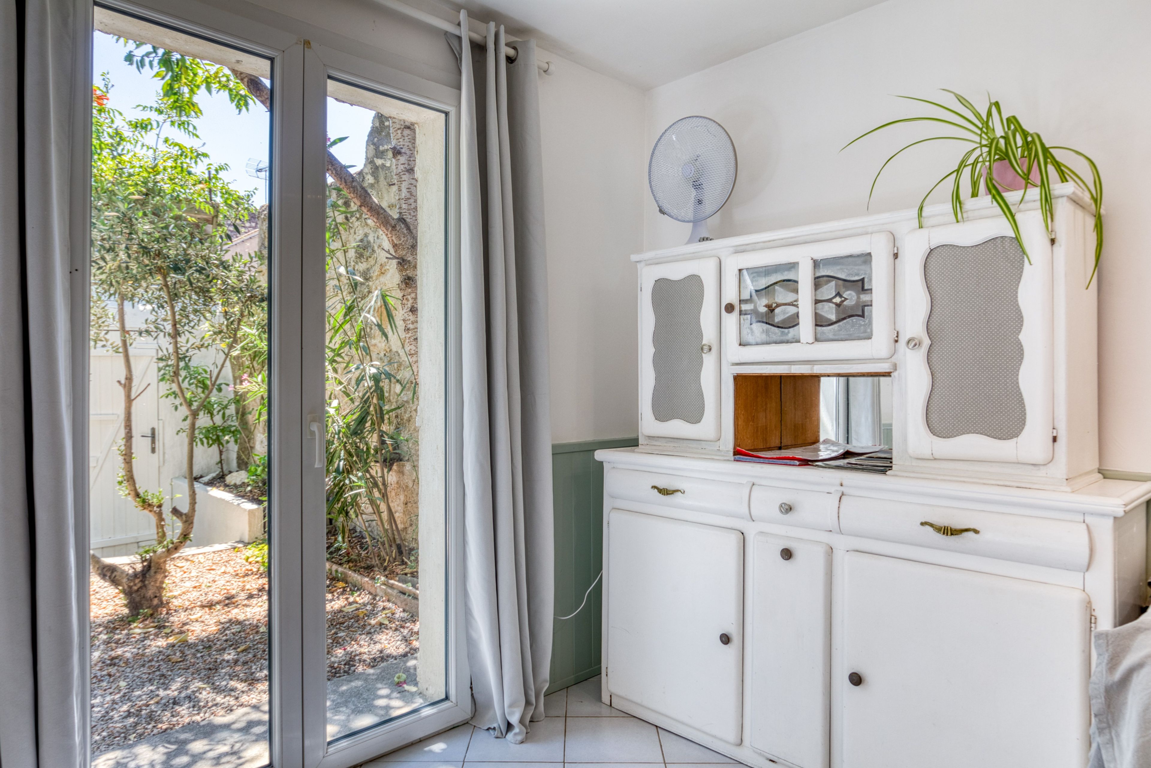 Ein Zimmer mit einer Schiebeglastür, die zu einem Garten führt, einem weißen Schrank mit Schubladen und einem Wandventilator.