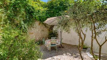 Un petit patio extérieur avec une table et des chaises, entouré d’une végétation luxuriante et d’un mur de pierre.