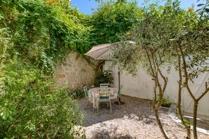 A small outdoor patio with a table and chairs, surrounded by dense foliage and a stone wall.
