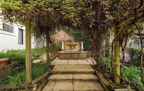 Un escalier en pierre mène à une fontaine située dans un jardin, avec un mur de briques et de la verdure en arrière-plan.