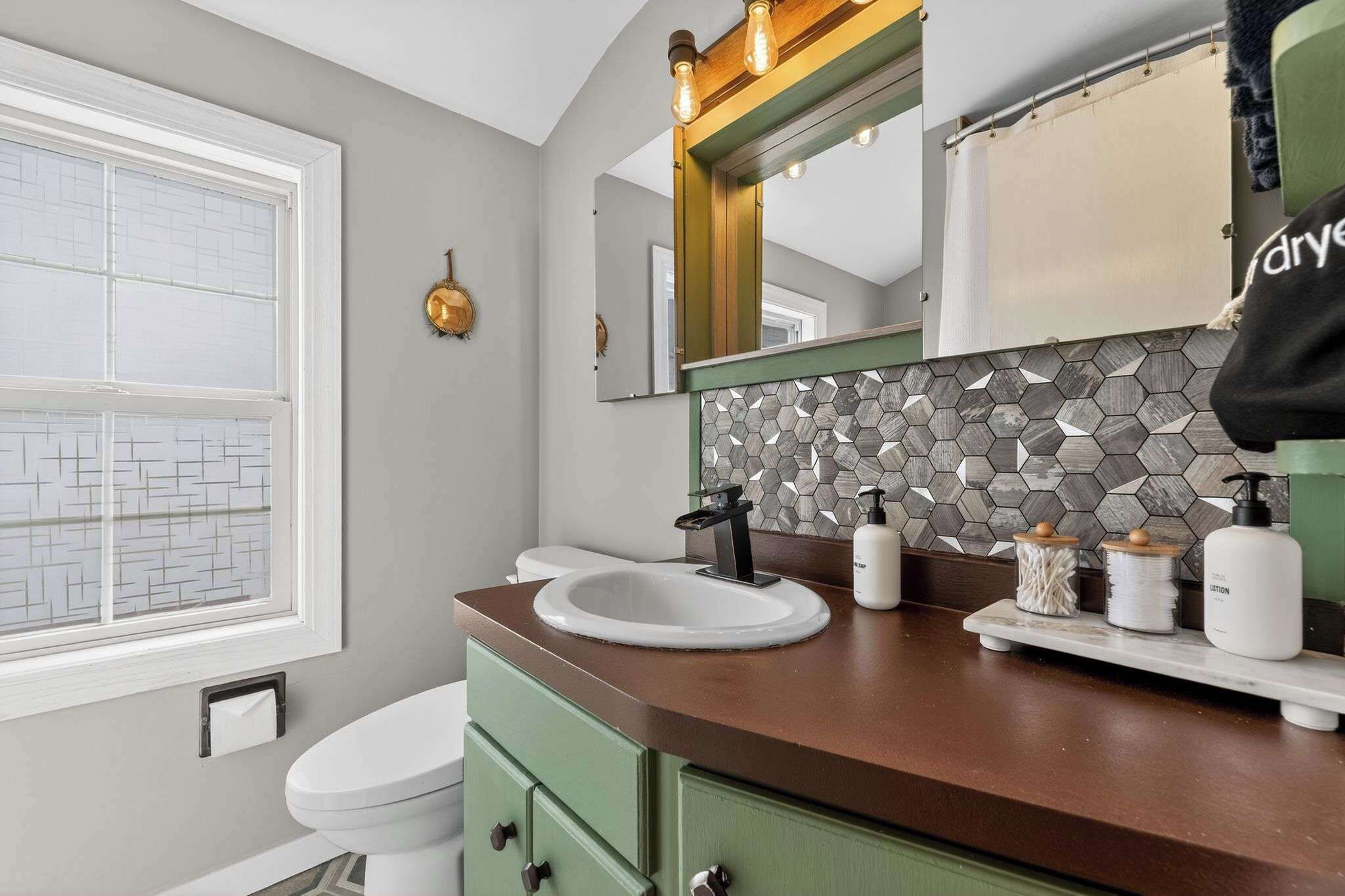 A bathroom with a white toilet, a round sink, a mirror, and a hexagonal tile backsplash.