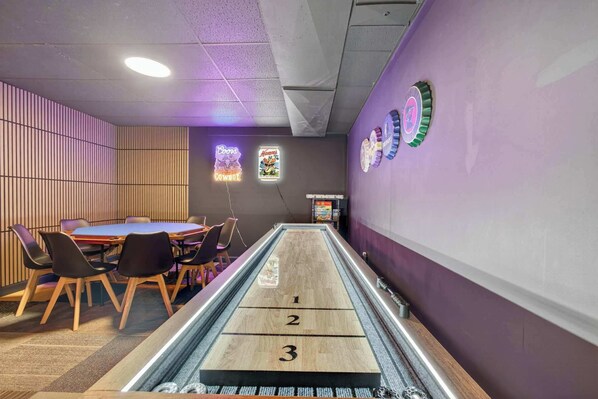 Ein Shuffleboard-Tisch mit nummerierten Enden, ein Essbereich mit StĂŒhlen und Tisch sowie eine Wand mit dekorativen Tellern.