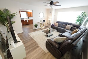 A living room with a brown leather sofa, a glass coffee table, a ceiling fan, and a kitchen in the background.
