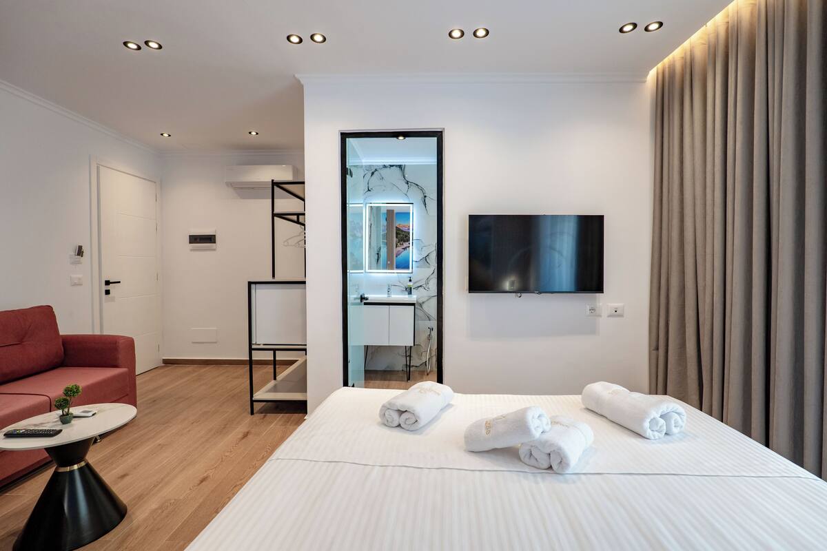A modern hotel room with a bed, a red sofa, a small table, and a TV mounted on the wall.