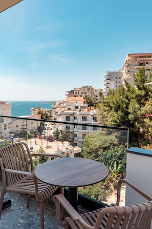 Un balcón con una mesa redonda y dos sillas, con vista a un paisaje urbano costero con edificios y árboles.