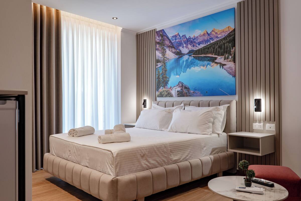 A hotel room with a large bed, a nightstand, a small table, and a painting of a mountain landscape.