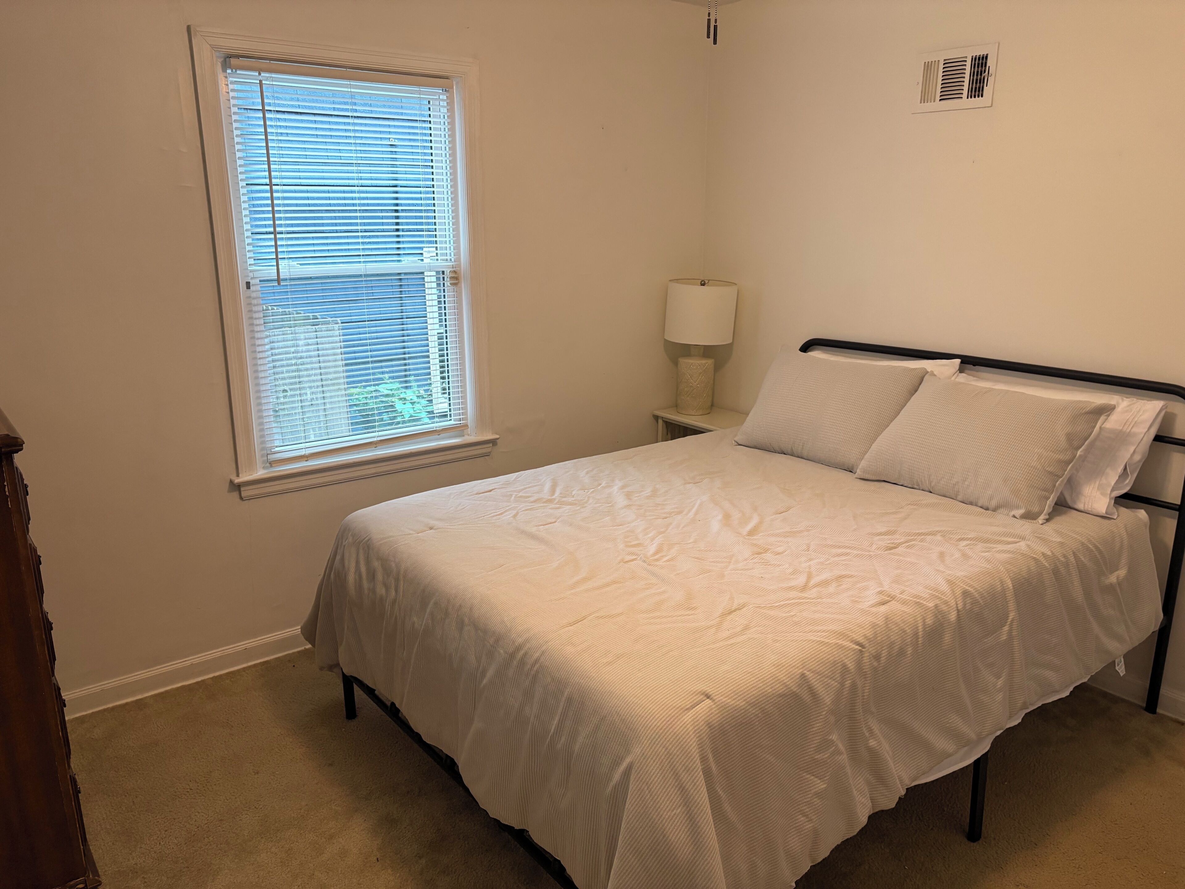 A bedroom with a bed, a window with blinds, a nightstand with a lamp, and a wall-mounted vent.