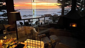 Un intérieur chaleureux avec vue sur l’océan au coucher du soleil, une cheminée et un coin repas avec des sièges à l’extérieur.