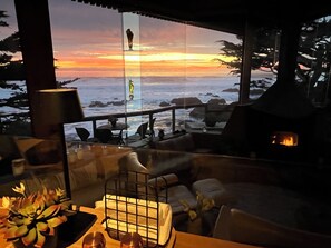 A cozy interior with a view of the ocean at sunset, a fireplace, and a dining area with outdoor seating.