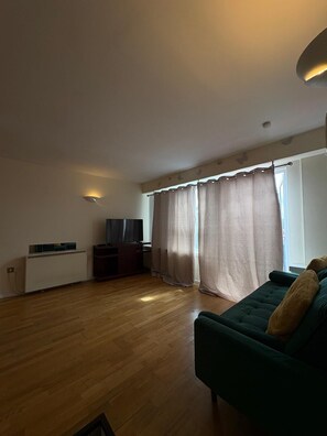 A living room with a green sofa, a TV, a radiator, and a window with curtains. - Beautiful 2 Bedroom - Woolwich (London)