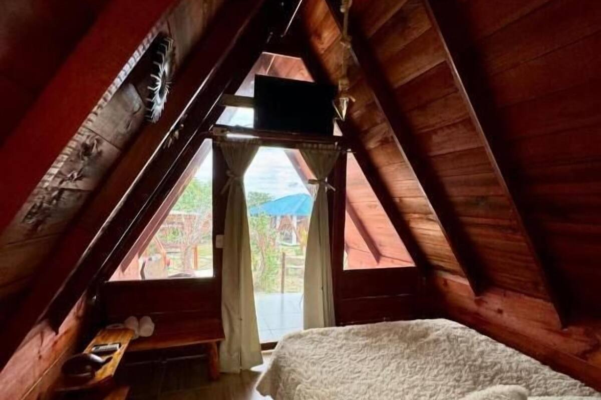 Una habitación con techo inclinado, una cama, un banco y vistas a través de una puerta.