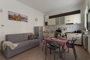 Eine moderne Küche mit weißen Schränken, einem Esstisch mit kariertem Tischläufer und einem grauen Sofa mit Decke.