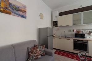 Eine kompakte Küche mit Kühlschrank, Mikrowelle, Backofen und Spüle. Im Wohnbereich befindet sich ein graues Sofa mit gemusterten Kissen.
