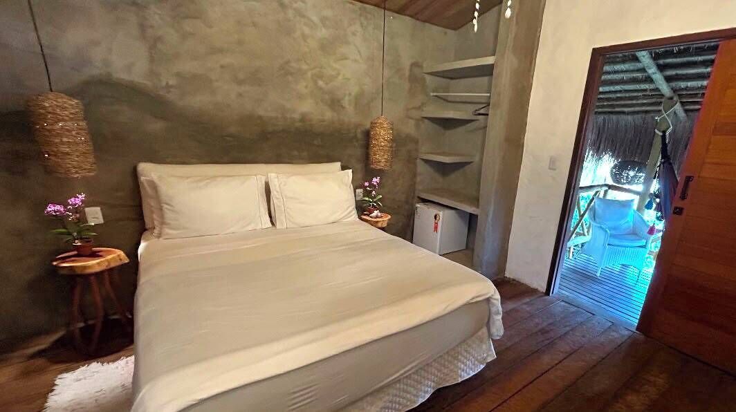 Un dormitorio con una cama, dos mesitas de noche, una lámpara colgante, una planta pequeña en maceta y una puerta que da a un espacio al aire libre.