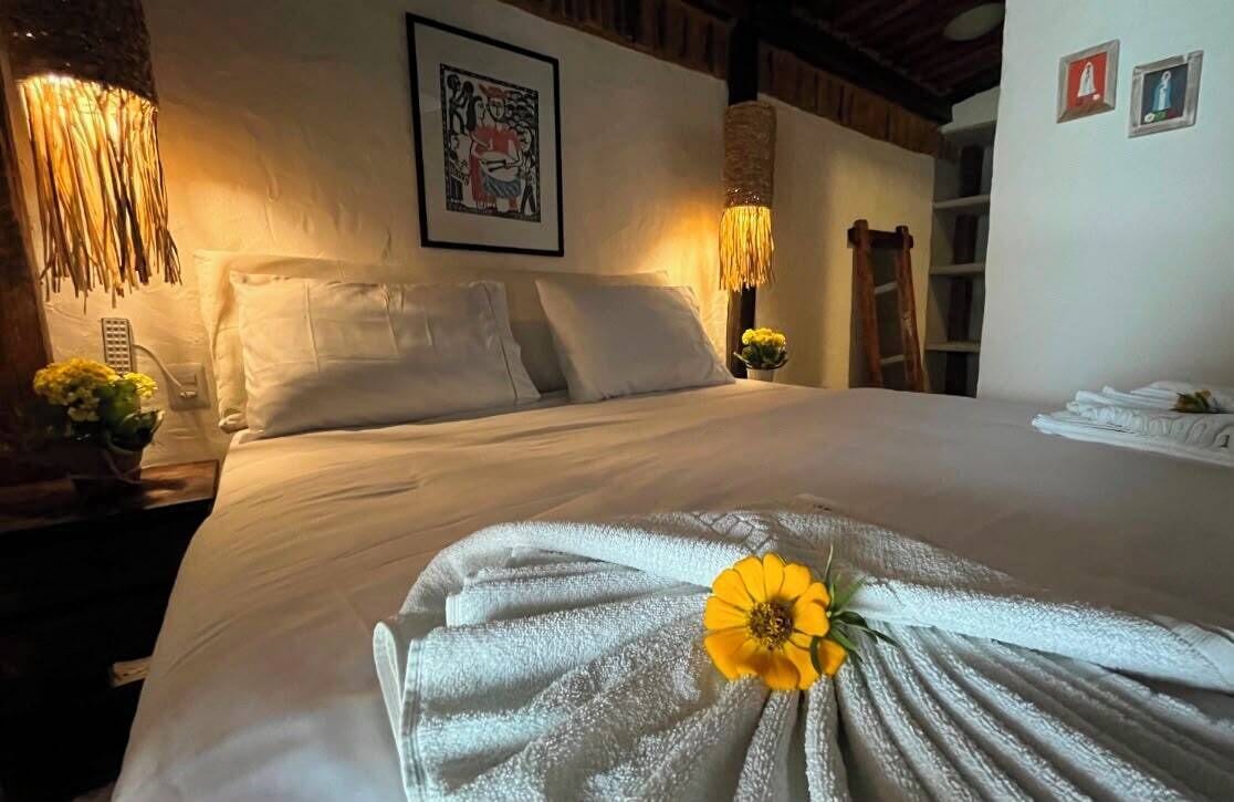 A neatly made bed with a towel and a flower, a wall-mounted picture, and a shelf with folded towels.