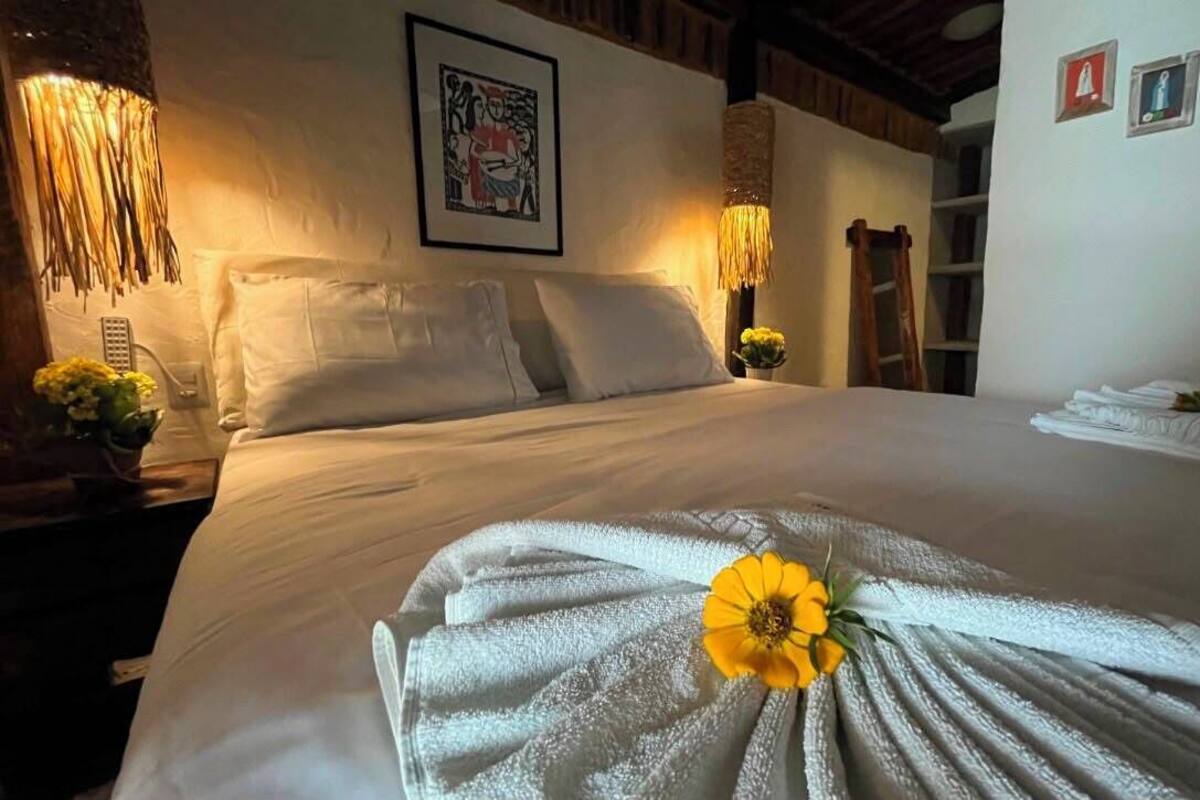 A neatly made bed with a towel and a flower, a wall-mounted picture, and a shelf with folded towels.