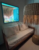 A room with a wooden sofa, a woven hanging light fixture, and a window with a view of a garden.