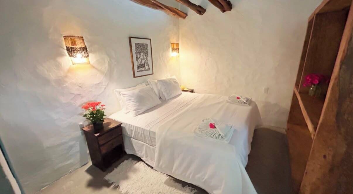 A bedroom with a bed, bedside tables, a nightstand, a wall-mounted lamp, and a shelf with flowers.