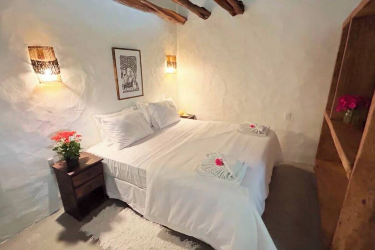 A bedroom with a bed, bedside tables, a nightstand, a wall-mounted lamp, and a shelf with flowers.