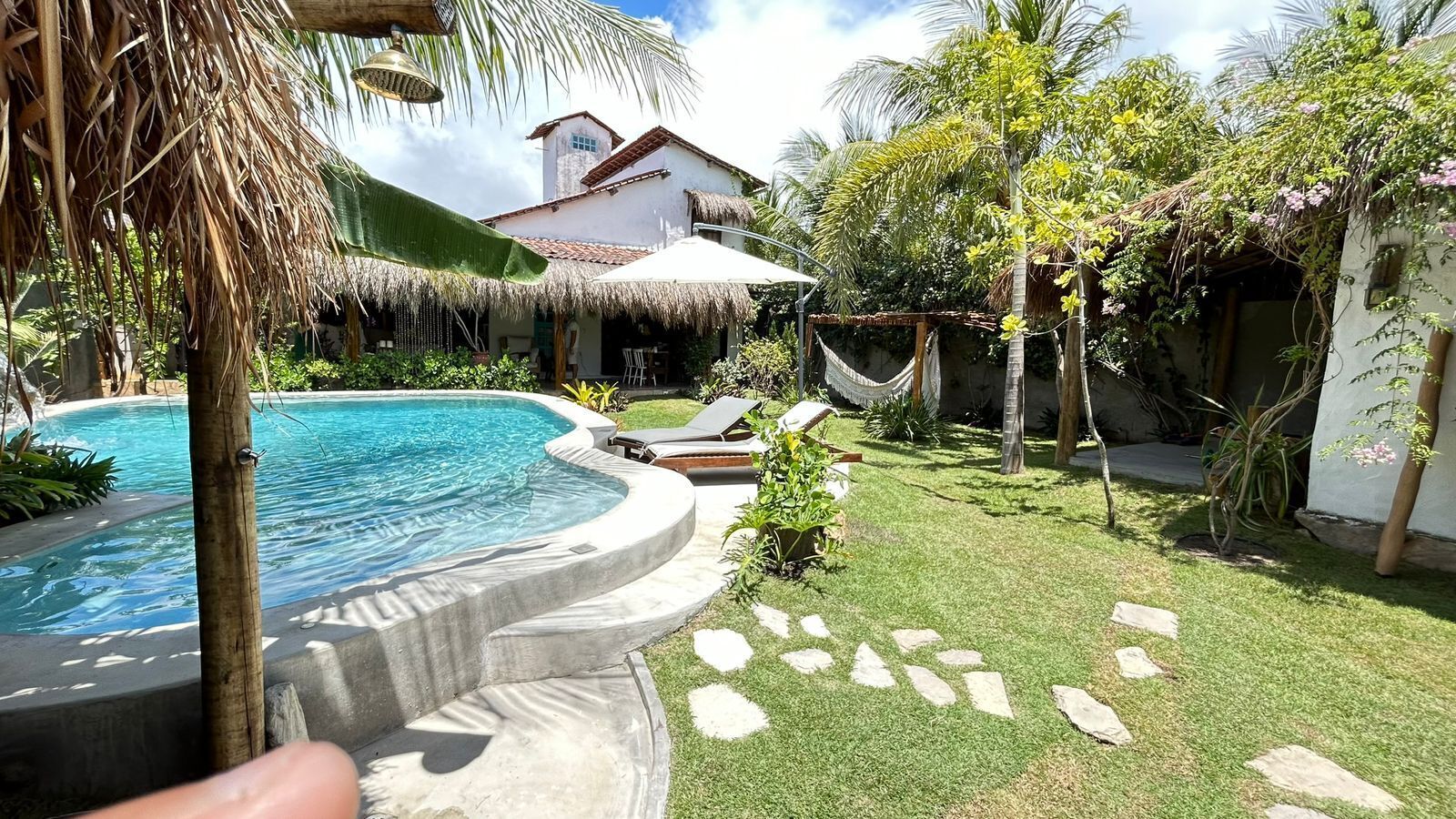 A swimming pool with a stone border, a thatched roof structure, and a white building in the background.