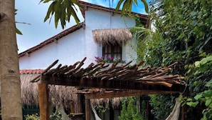Eine Hängematte unter einer Holzpergola in einem üppigen Garten, im Hintergrund ein weißes Gebäude.