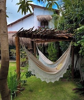 A hammock hanging under a wooden pergola in a lush garden with a white building in the background.
