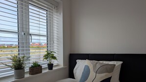Quarto com cama, janela com persianas e duas plantas em vasos no parapeito.