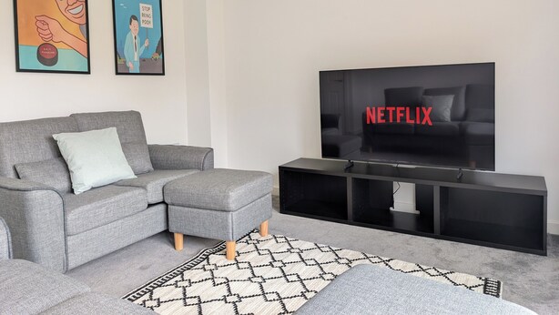 Uma sala de estar moderna com um sofá cinzento, um móvel para entretenimento preto e uma TV exibindo 'NETFLIX'.