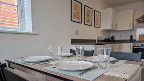 Mesa de jantar posta com pratos, copos e talheres, com uma cozinha ao fundo.