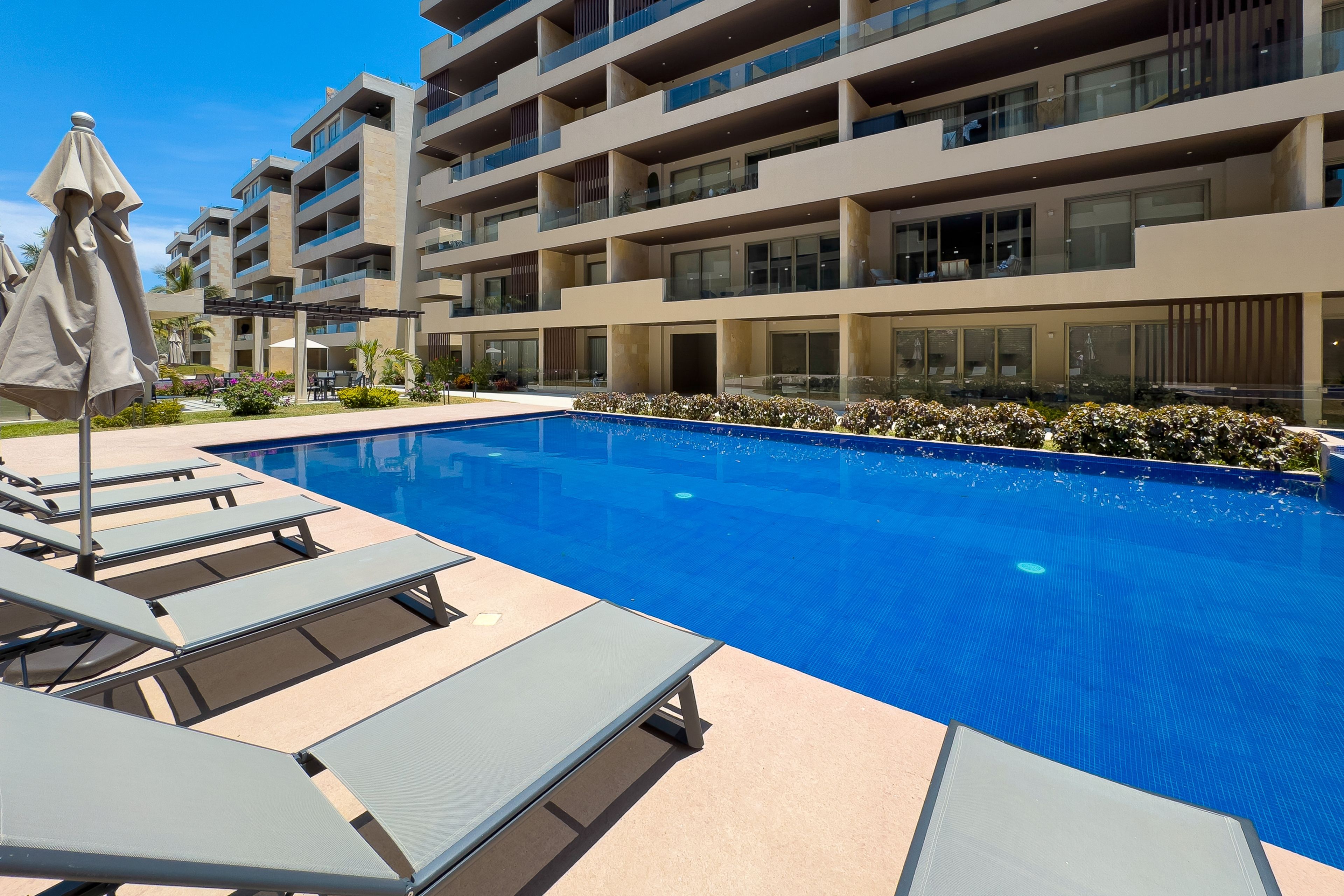 A swimming pool with lounge chairs and umbrellas, surrounded by a multi-story building with balconies.