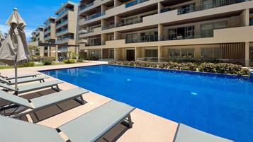 A swimming pool with lounge chairs and umbrellas, surrounded by a multi-story building with balconies.