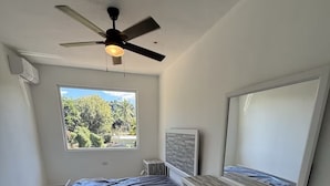 Ein Schlafzimmer mit Bett, Deckenventilator, Fenster mit Aussicht, Kommode und Spiegel.