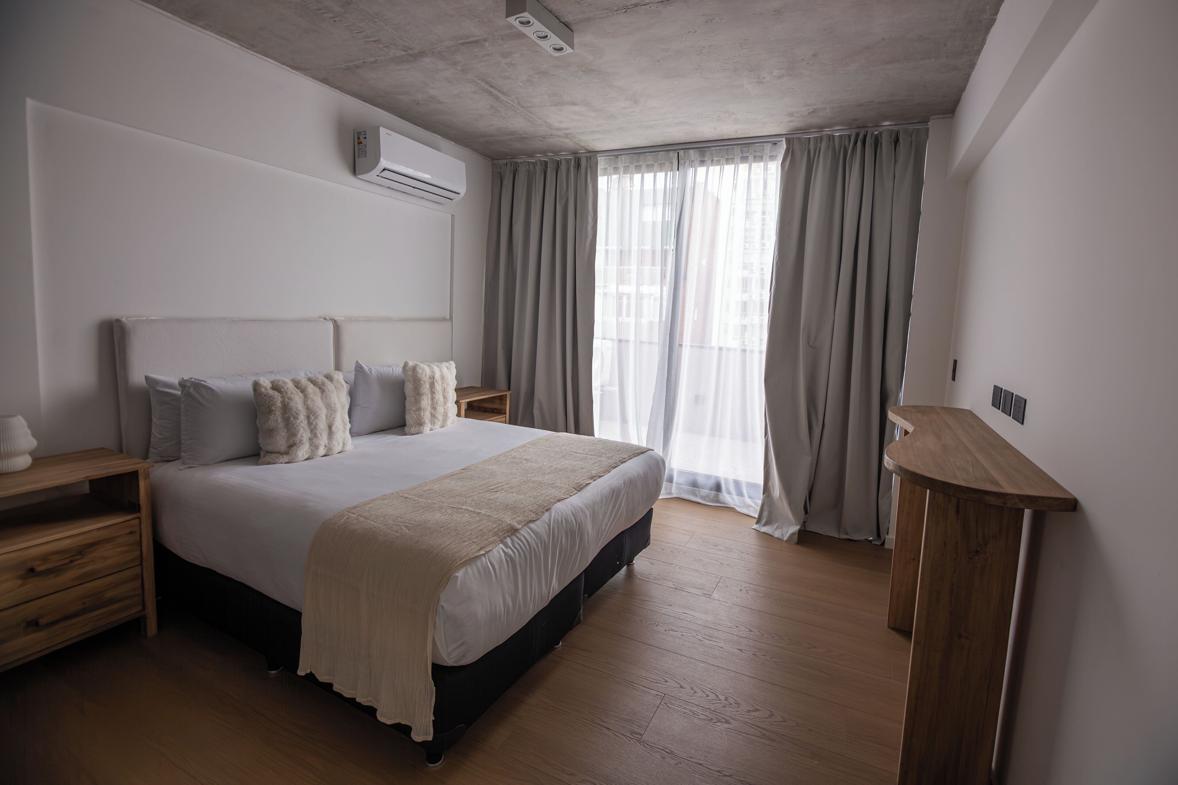 Ein Schlafzimmer mit einem Bett, Nachttischen aus Holz, einem Beistelltisch, einer Holzkommode und einem Fenster mit Vorhängen.